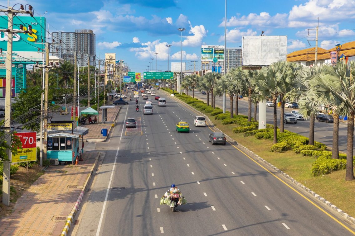 Große Straße in Pattaya, Thailand, mit Autos und Motorrädern auf beiden Fahrspuren, gesäumt von Palmen und Gebäuden im Hintergrund