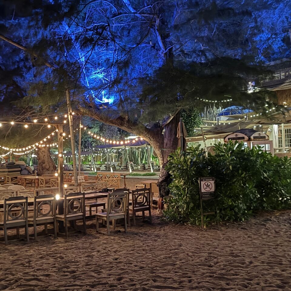 Strandrestaurant bei Nacht Sitzbereich am Strand mit Tischen, Stühlen, Lichterketten und Bäumen bei Nacht